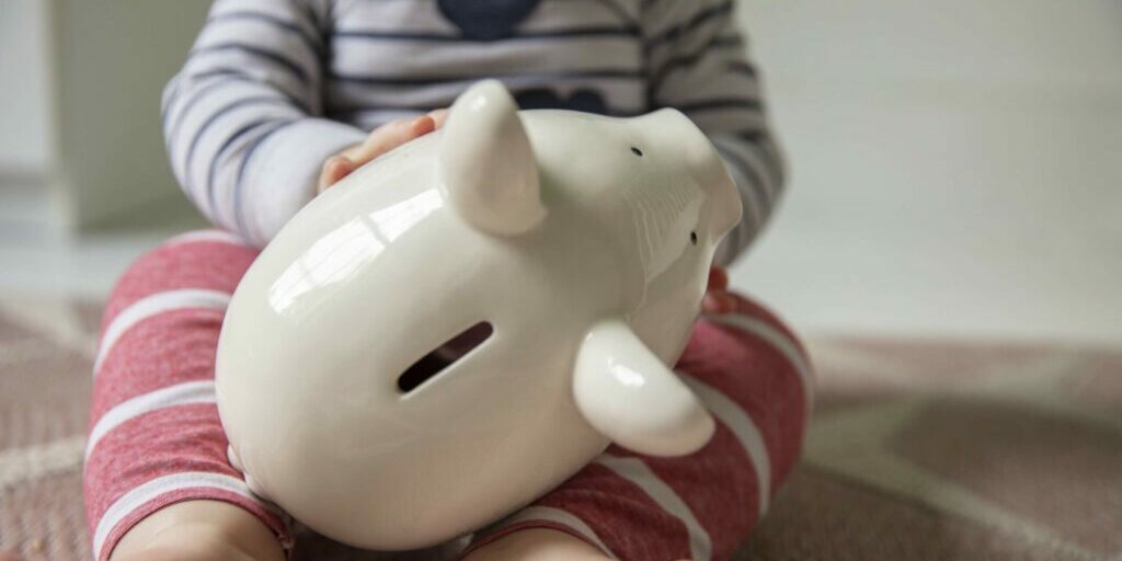 Cost of childcare concept. Young baby holds onto a white piggy bank